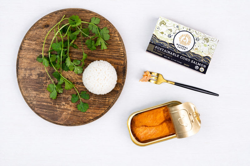 Natural Catch Coho Salmon in Teriyaki Sauce flat lay: opened tin, carton, fork and a scoop of rice with cilantro on a wood board