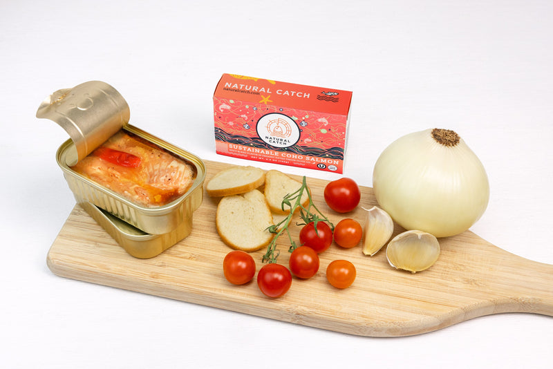 Natural Catch Coho Salmon Spicy Blend tin and current coral package on a cutting board with cherry tomatoes, garlic, onion, and bread