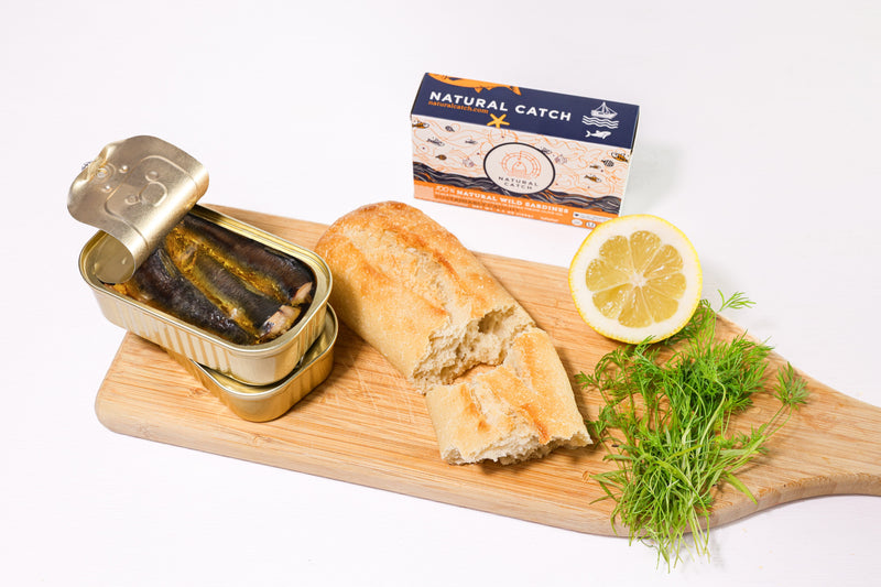Open can of Natural Catch Sardines on a board with baguette, dill and lemon