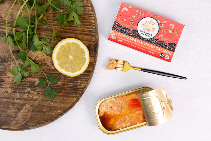 Opened tin of Natural Catch Coho Salmon Spicy Blend on a wood board with parsley, lemon, and the current coral package