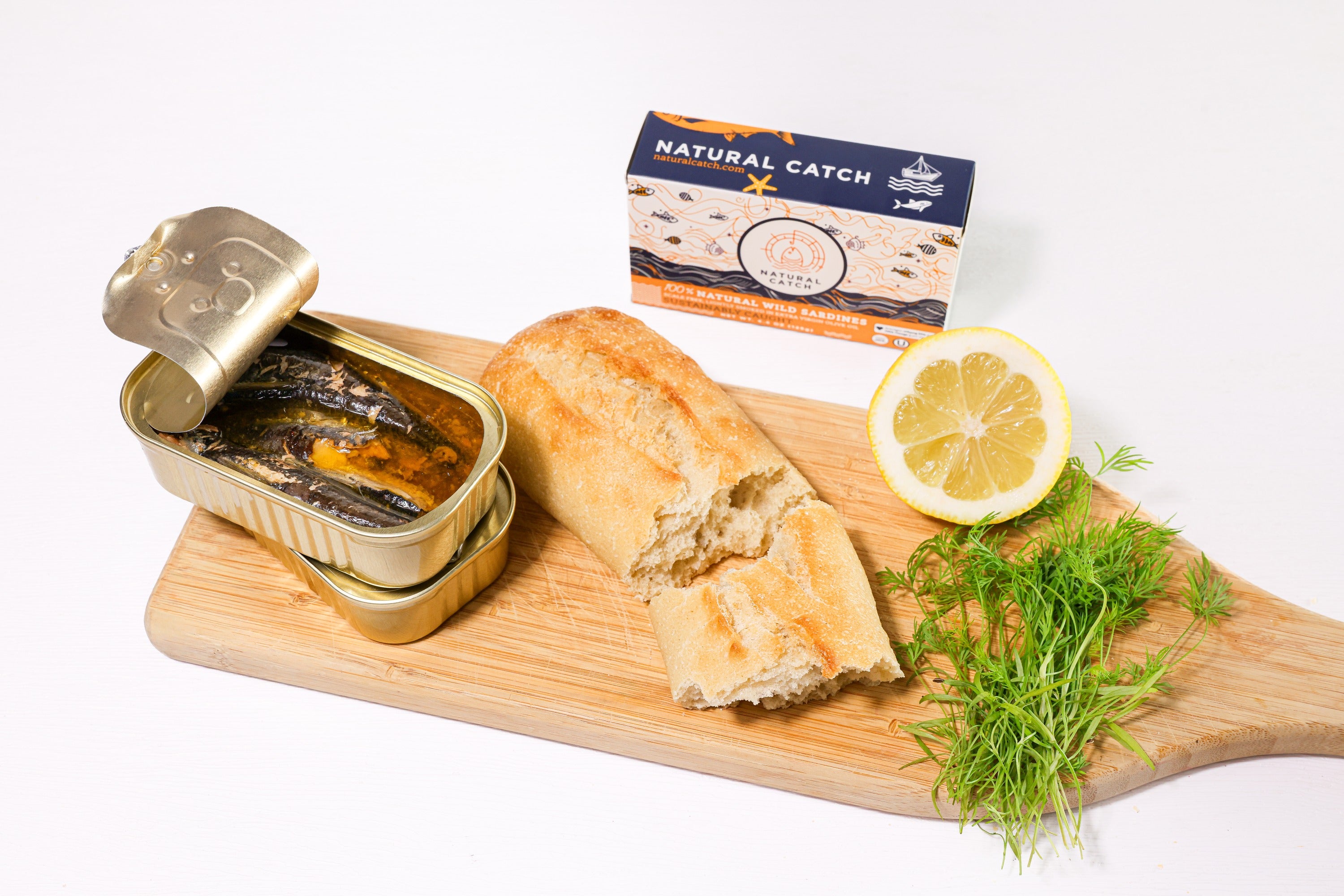 Open can of Natural Catch Sardines with baguette, dill and lemon on a wood board