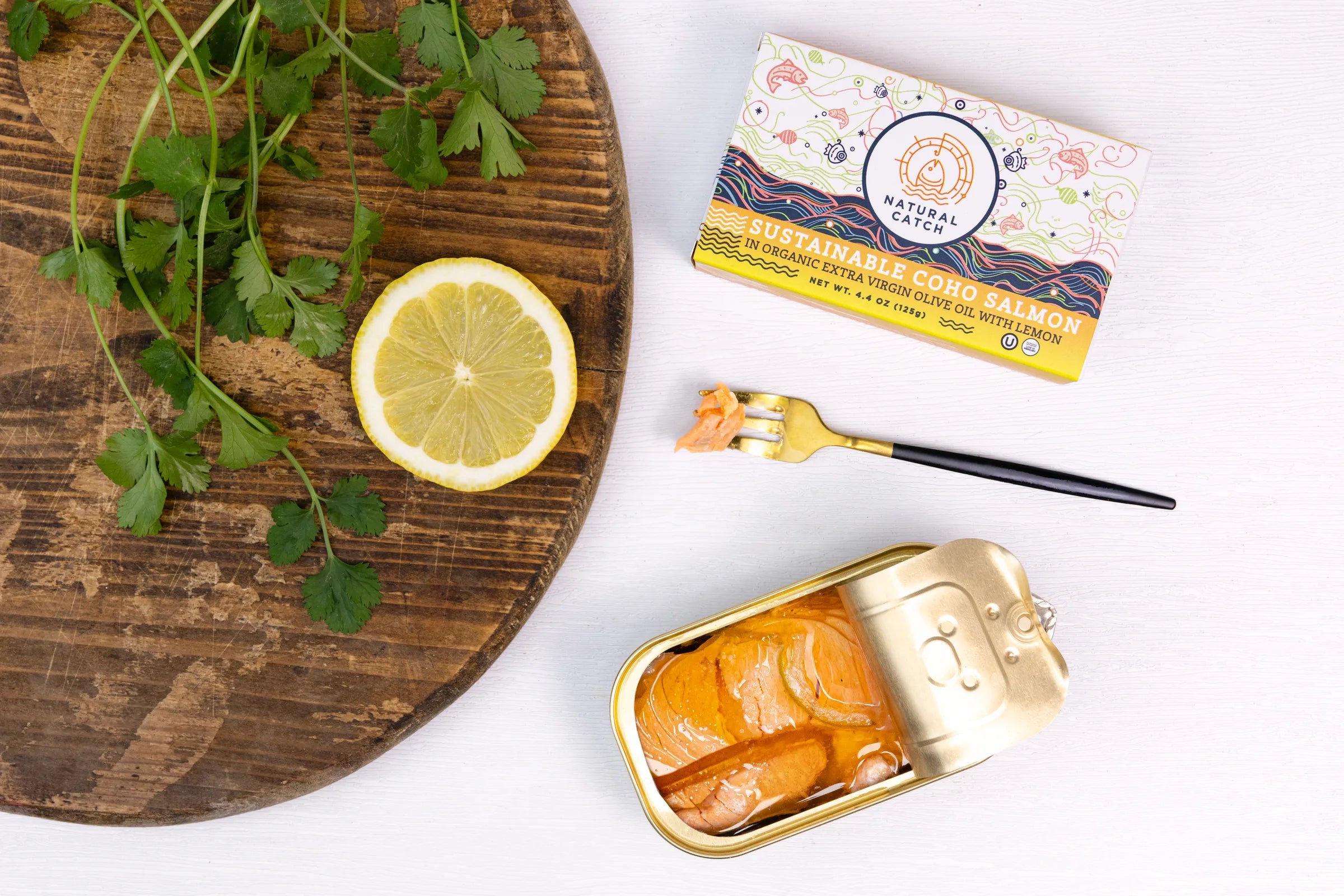 Opened tin of Natural Catch Coho salmon on wooden board with fresh lemon, cilantro, and product packaging